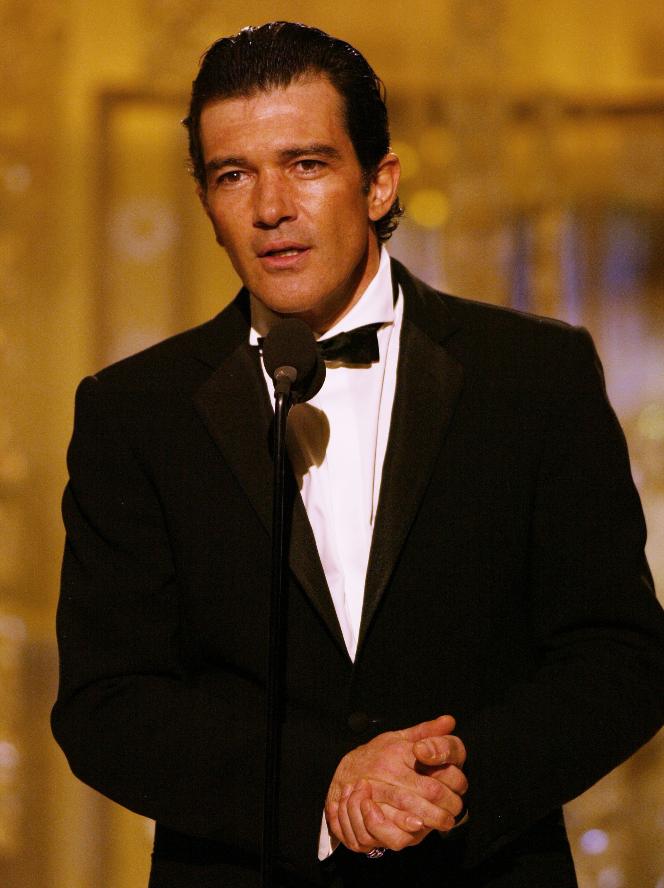 Antonio Banderas stands at a microphone in a tuxedo with a bow tie, speaking at an event