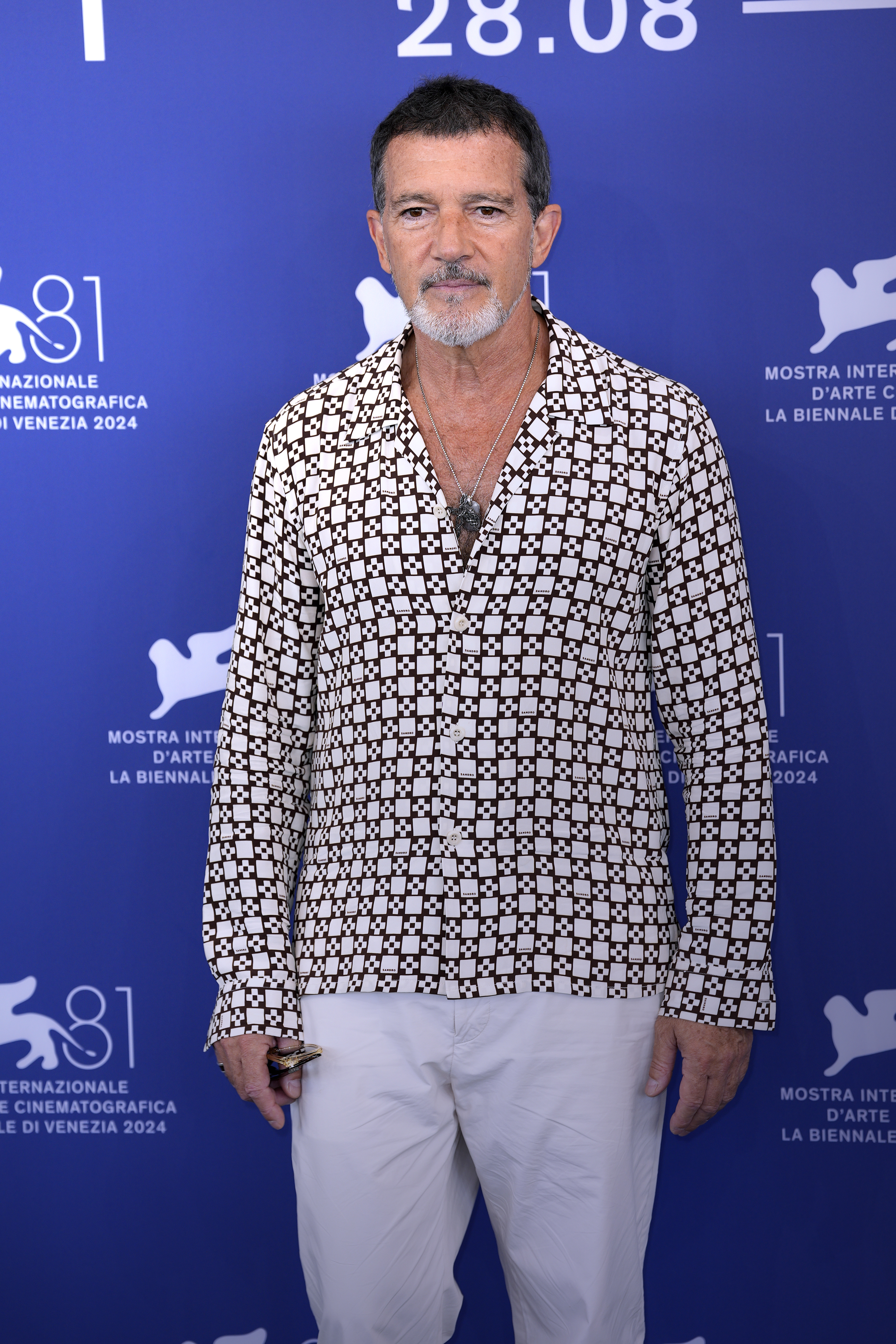 Antonio Banderas stands in front of a backdrop at the Venice International Film Festival, wearing a patterned shirt and white pants