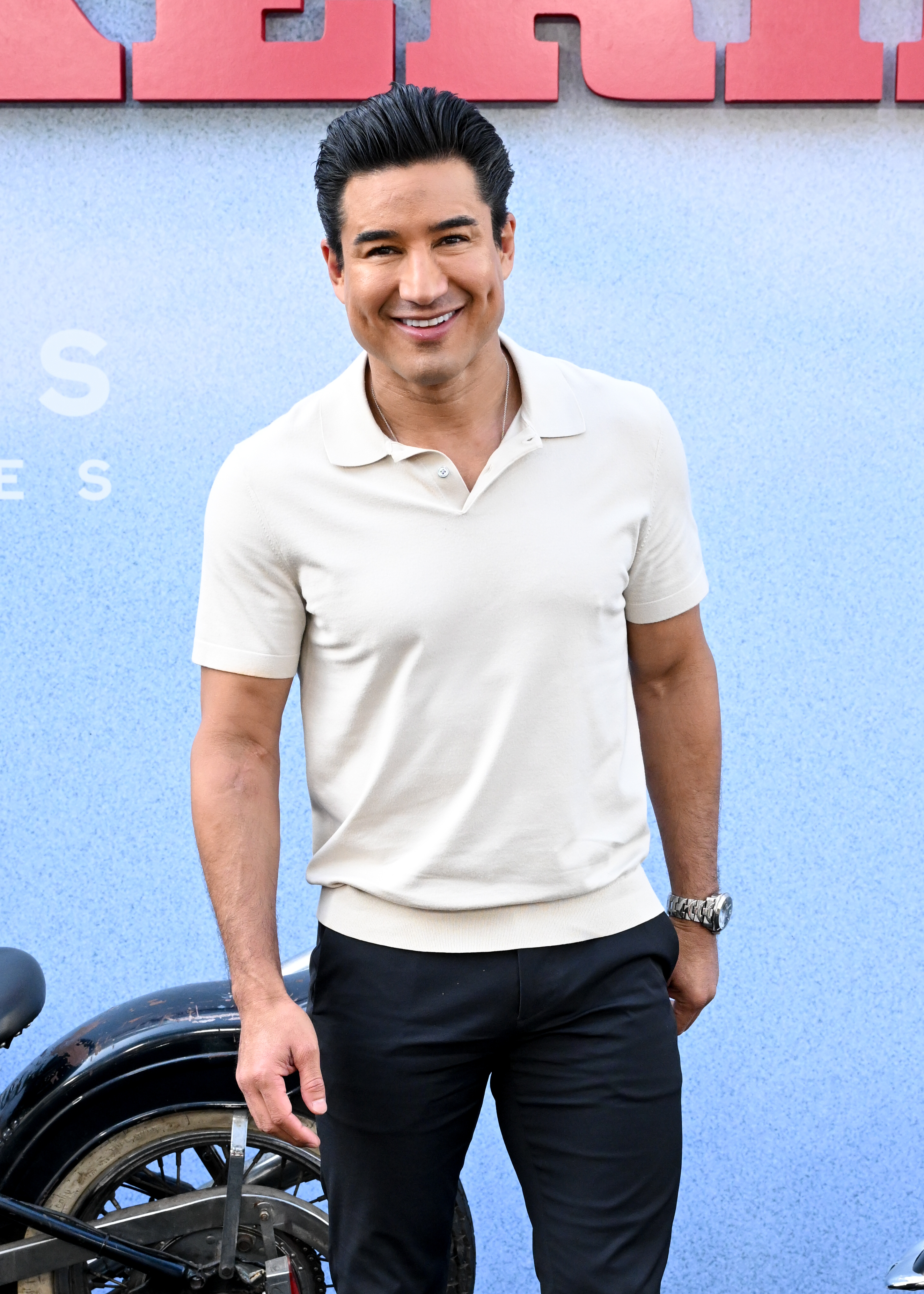 Mario Lopez smiles casually in a light polo shirt and dark pants, posing in front of a motorcycle at a public event