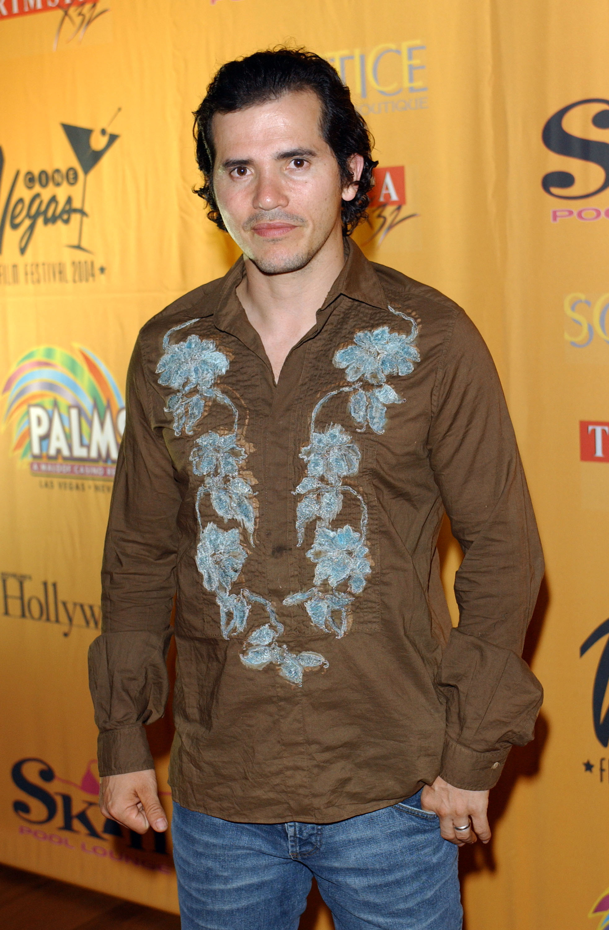 John Leguizamo at a red carpet event, wearing a brown shirt with blue floral embroidery and jeans