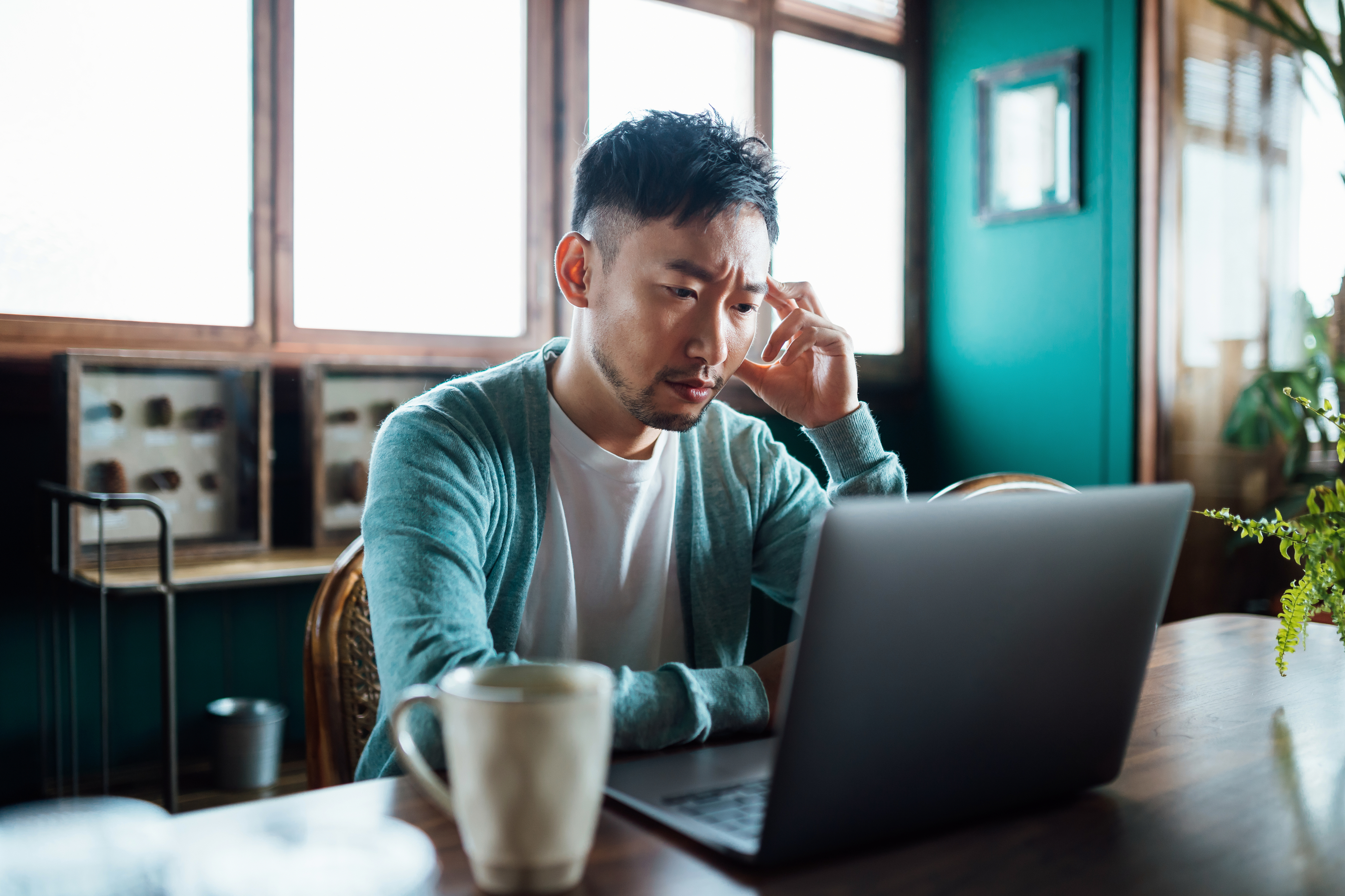 A man in a casual outfit sits at a table, looking pensive while using a laptop, with a coffee mug in front of him. The setting is a cozy living space
