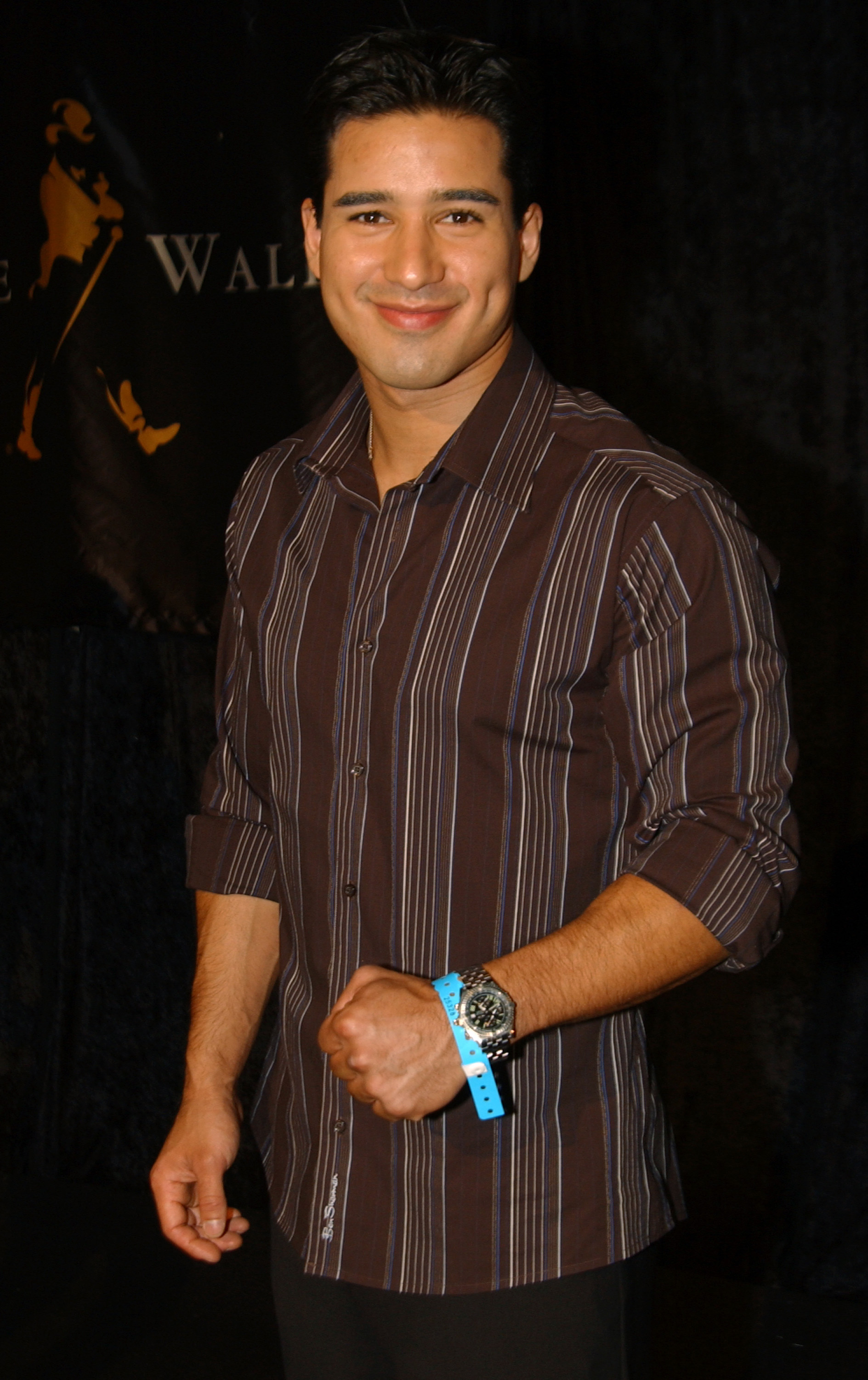 Mario Lopez smiling, wearing a striped button-up shirt, a wristband, and a watch