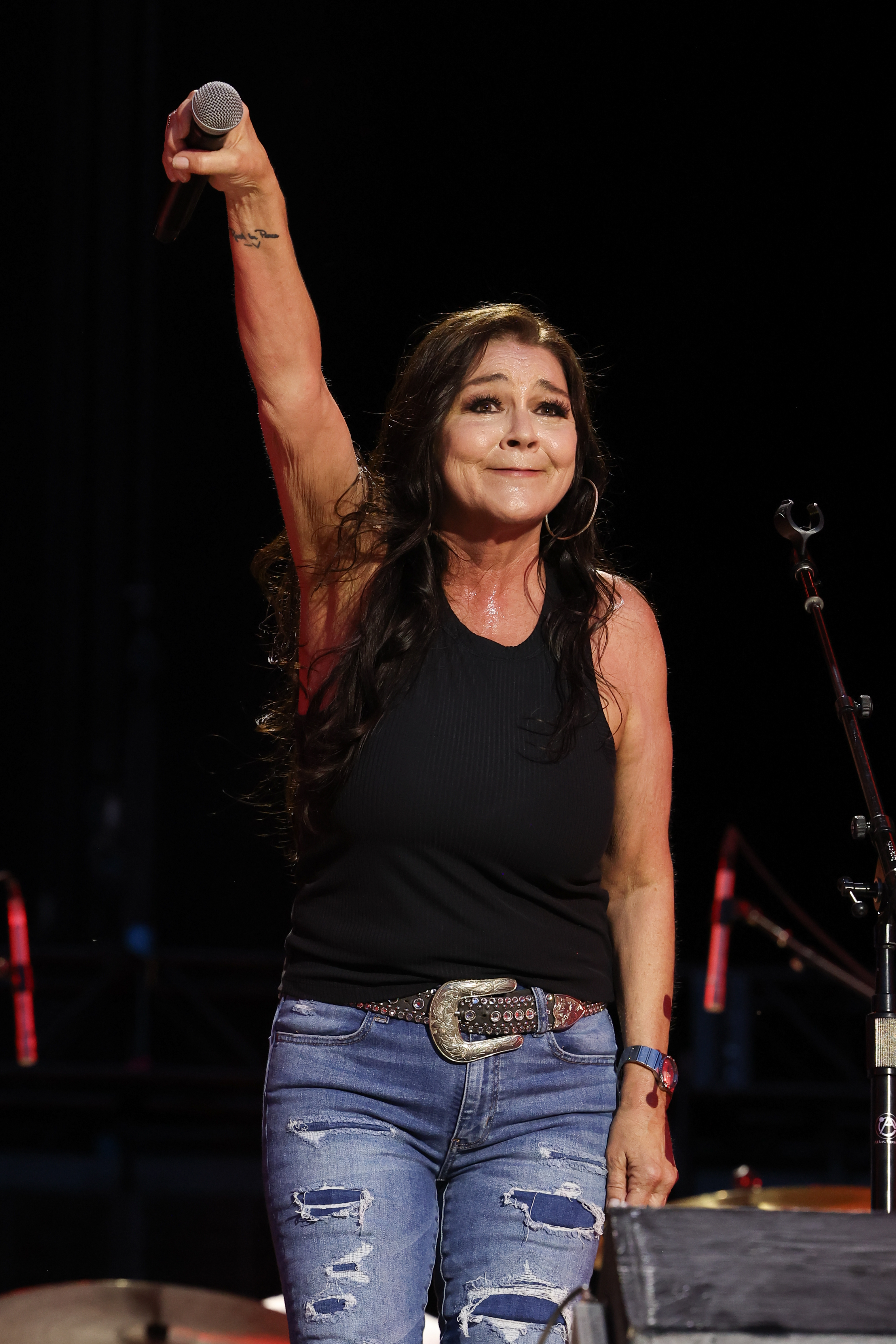 A woman in a sleeveless top and jeans, identified as a celebrity, holds a microphone while performing on stage