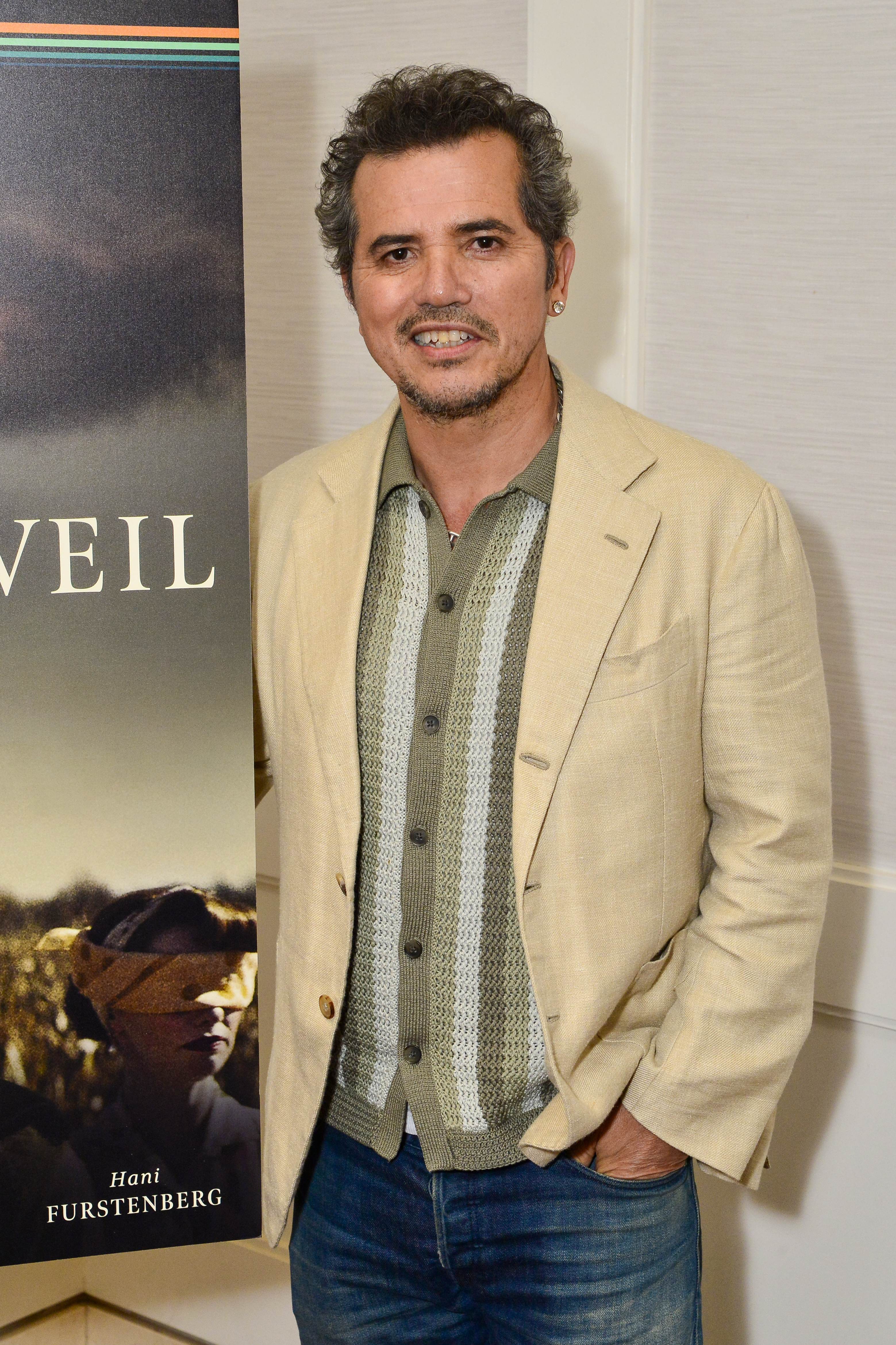 John Leguizamo poses in a casual outfit with a beige blazer and a patterned shirt at an event, standing next to a "Furstenberg" sign