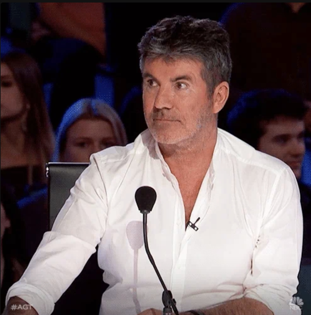 Simon Cowell, wearing a white shirt, is seated at a judge's panel with a serious expression, speaking into a microphone during a TV show