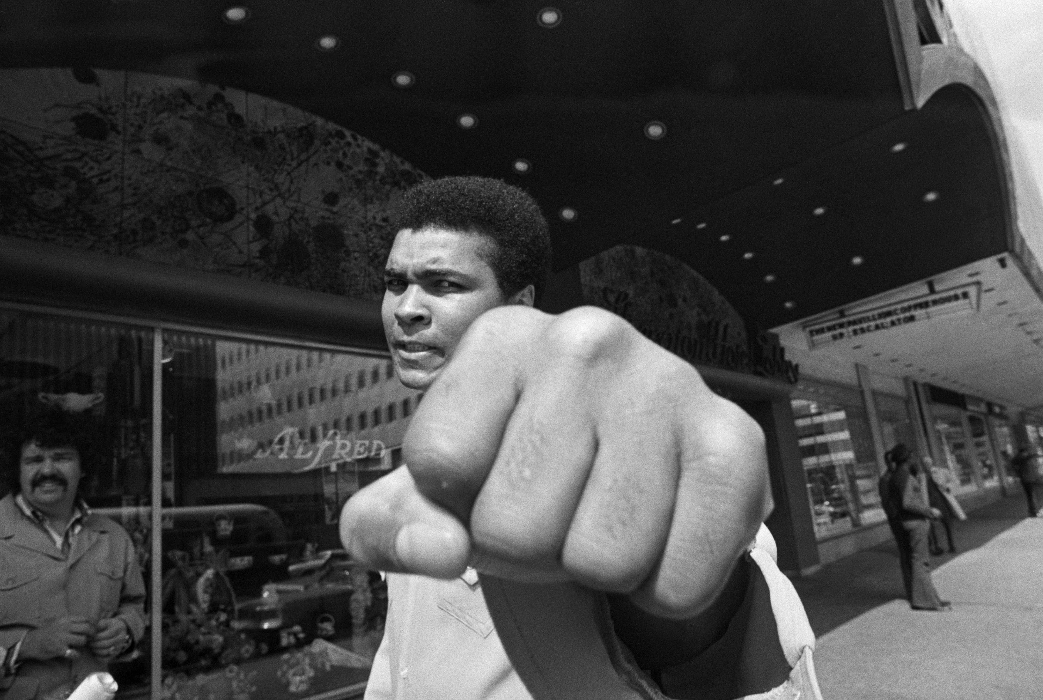 Muhammad Ali poses with his fist extended towards the camera on a city street