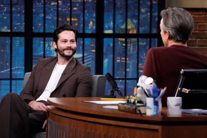 Dylan O'Brien in conversation with talk show host Seth Meyers on a late-night talk show set with cityscape backdrop