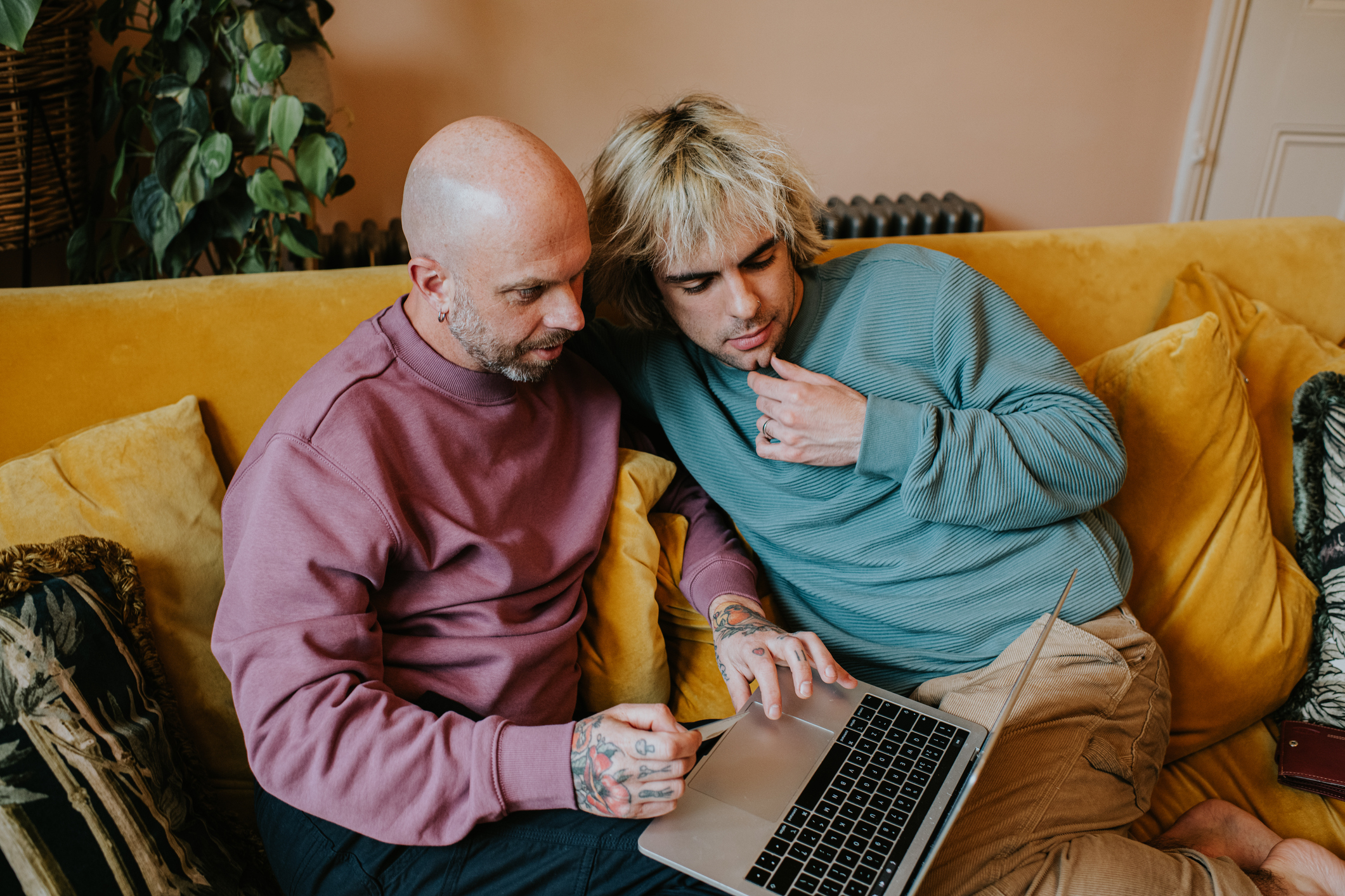 Two men, one bald with tattoos and in a sweatshirt, and the other with blonde hair in a long-sleeve shirt, sit on a couch looking at a laptop
