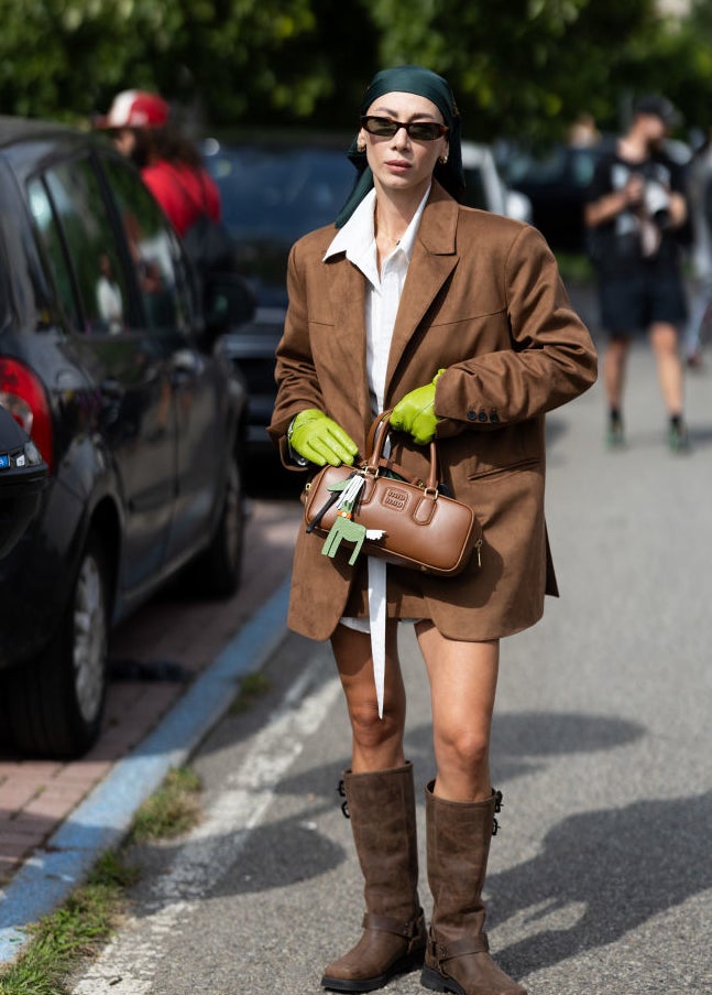 An individual wearing an oversized brown blazer, white shirt, green gloves, black headscarf, sunglasses, and brown boots is holding a bag on a street