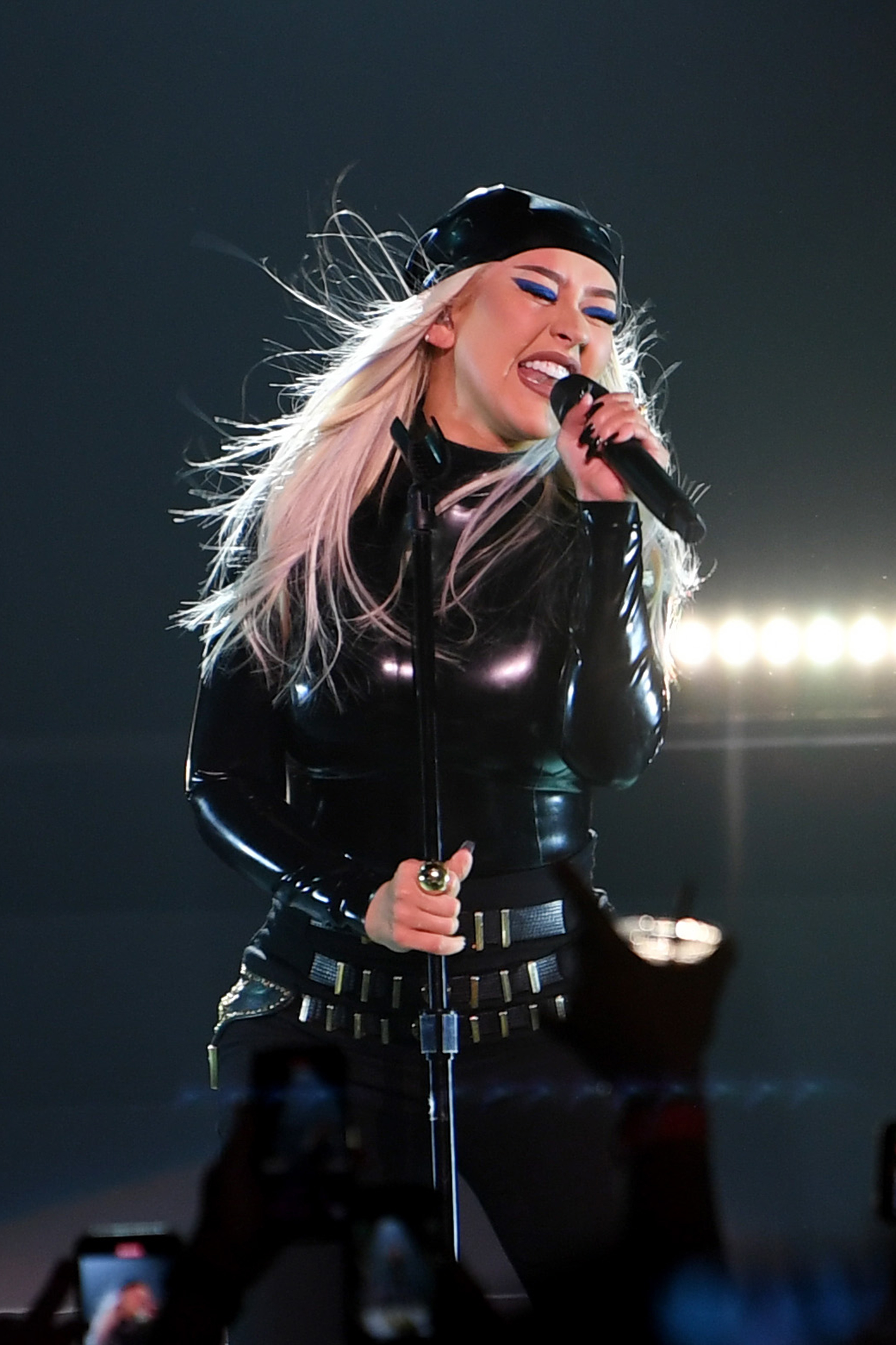 Christina Aguilera performs on stage, wearing a black leather outfit and cap, singing passionately into a microphone