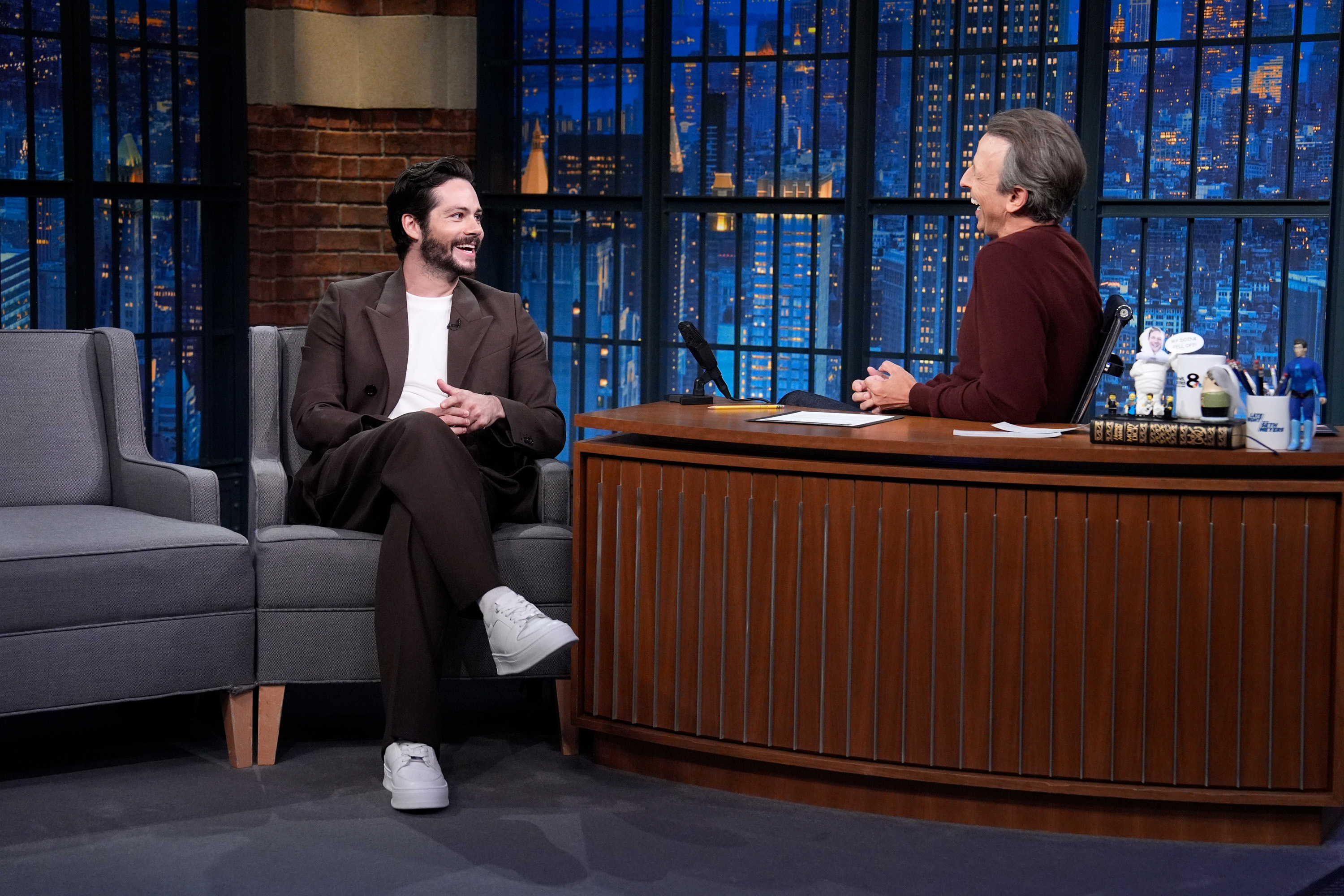 Dylan O'Brien, in a suit with sneakers, on a talk show set talking and smiling with Seth Meyers behind a desk