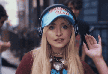 A woman wearing headphones and a cap with "Baby Girl" text on it dances to music on a street