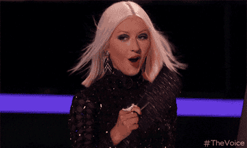 Christina Aguilera on The Voice, holding a fan with a surprise expression, wearing a stylish, black, sequined outfit with bold earrings