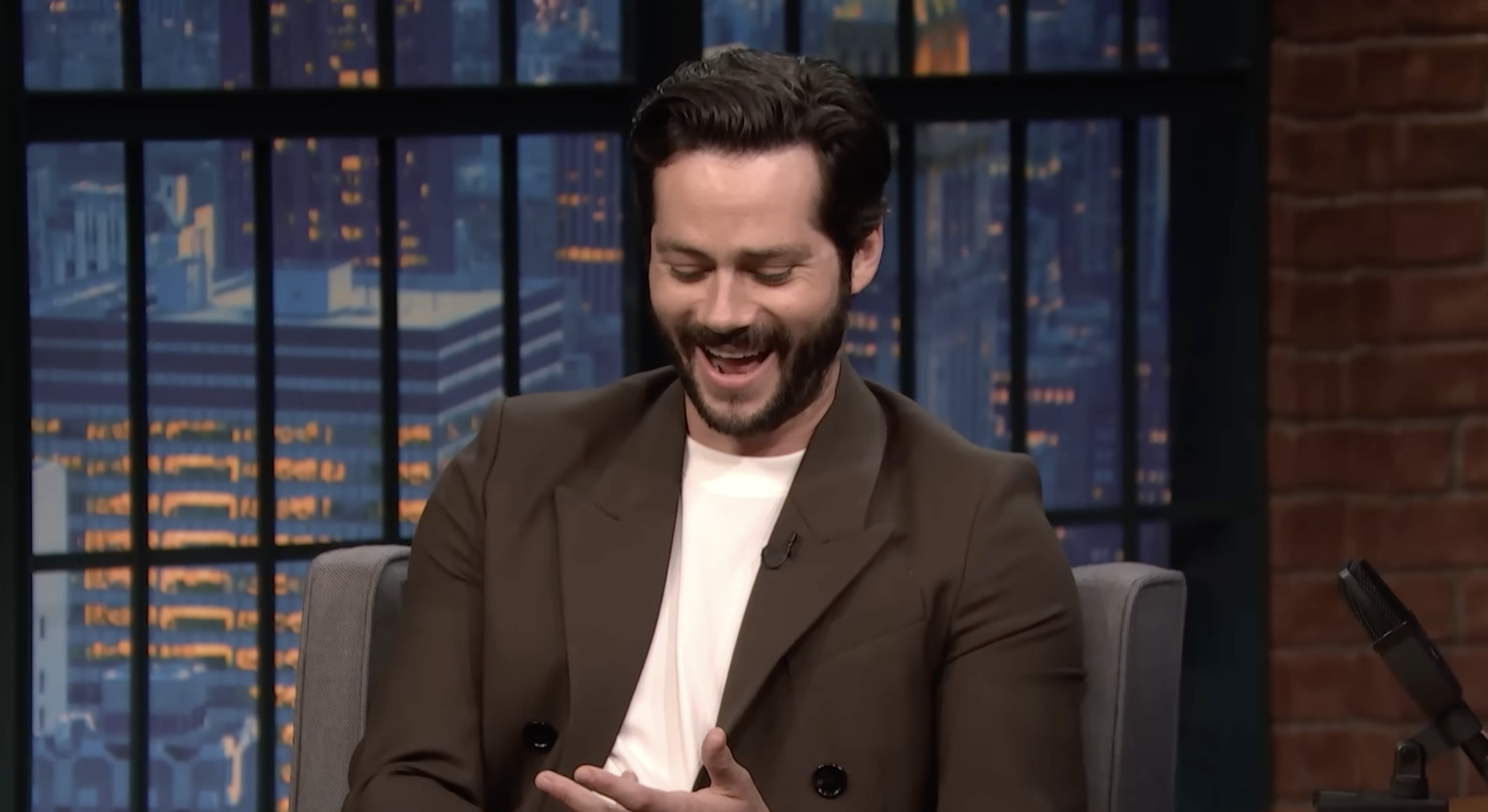 A man in a dark suit and white shirt laughs while sitting on a talk show set. A cityscape is visible through the window behind him