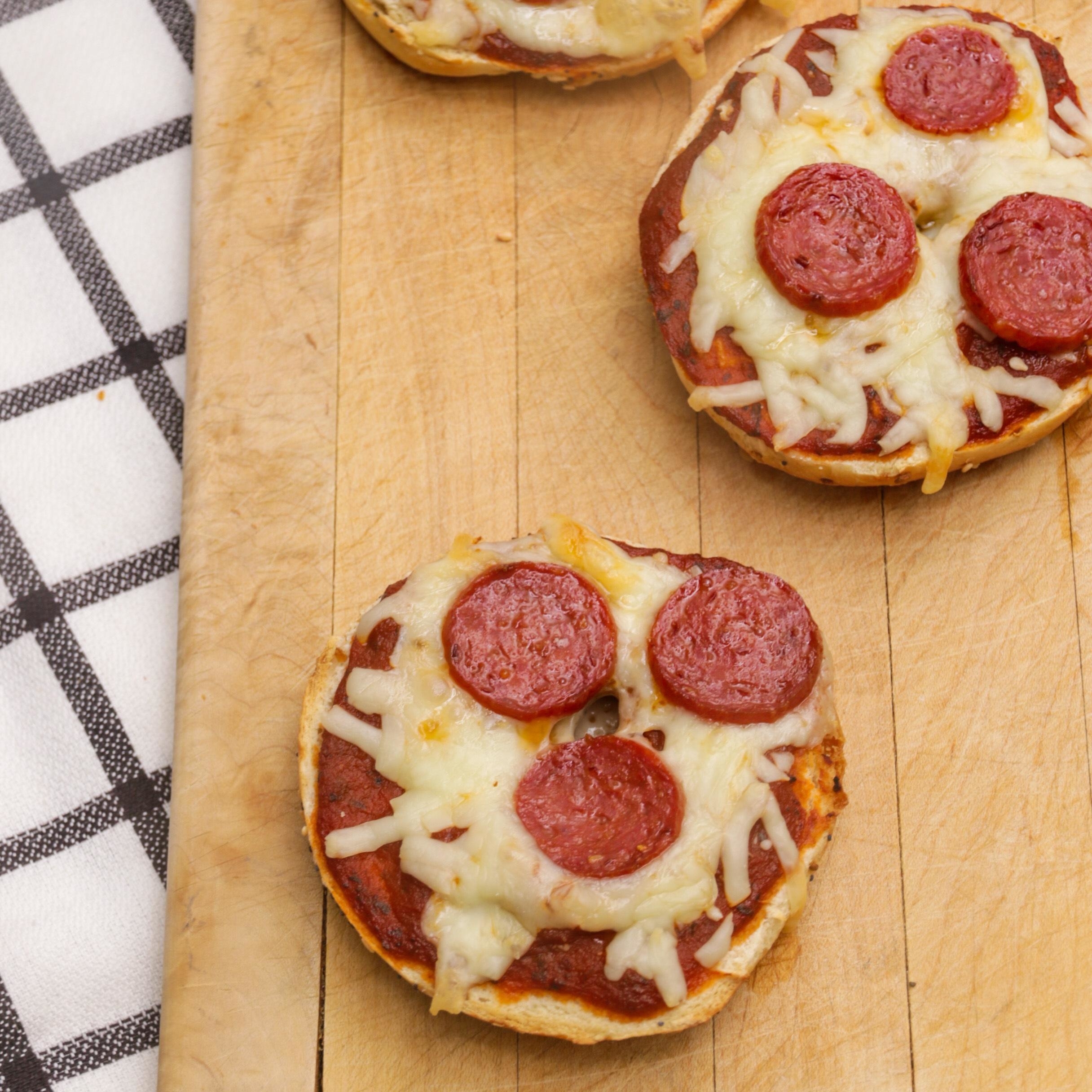 Mini pizza bagels topped with melted cheese and pepperoni slices placed on a wooden board next to a checkered napkin