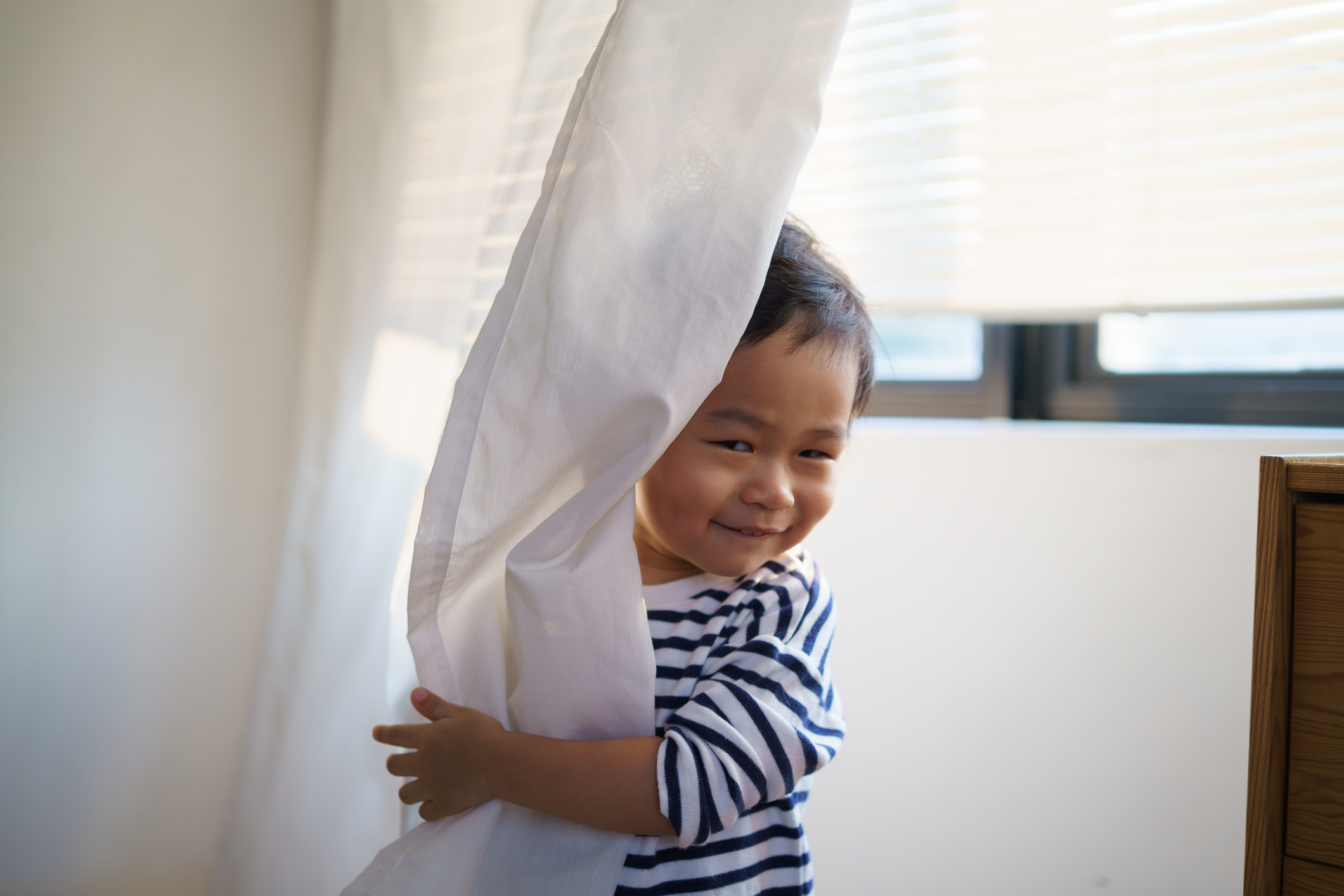 A toddler with short hair and a striped shirt peeks out playfully from behind a curtain in a sunlit room