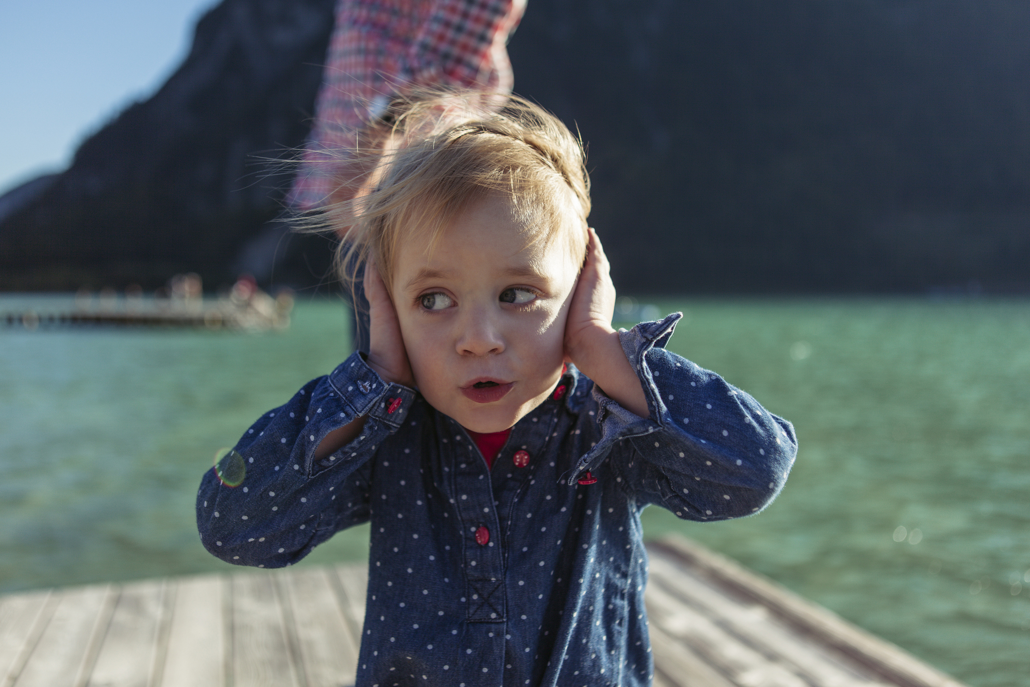 A child with blonde hair, wearing a patterned blue shirt, stands covering their ears by a lake. An adult stands blurred in the background