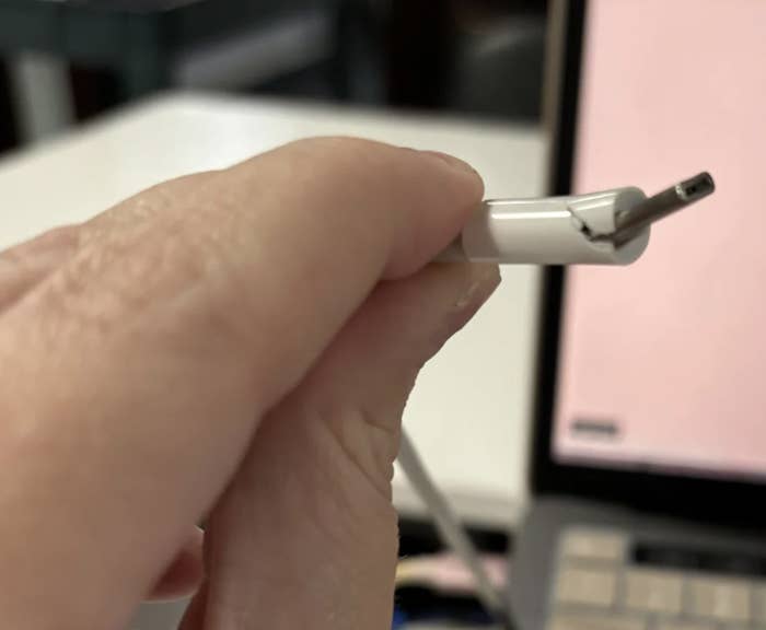 Close-up of a hand holding a frayed and damaged charging cable, with a laptop in the background