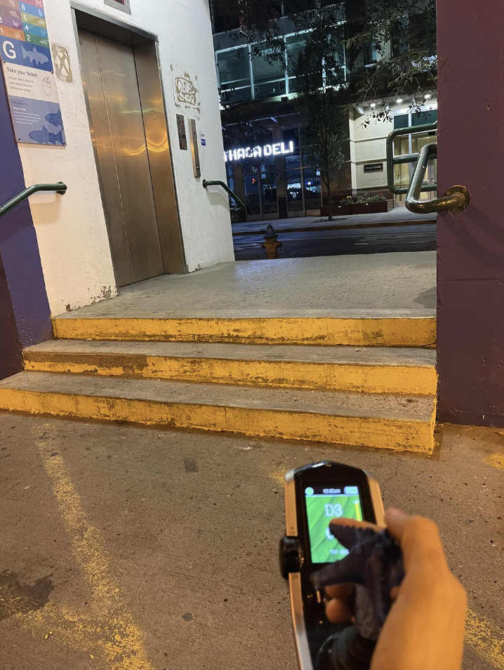 A person holds a device displaying "D3" in front of a flight of stairs leading to a building entrance, with Ghirardelli sign visible in the background