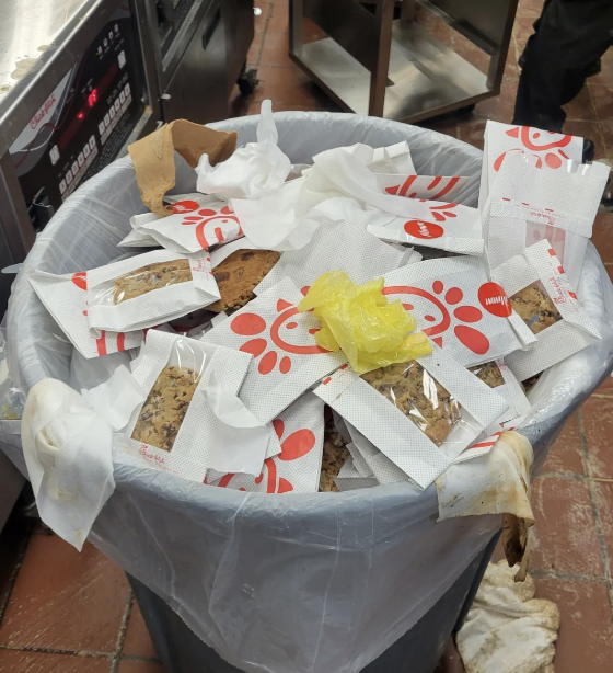 Trash can filled with numerous discarded Chick-fil-A cookie wrappers, tissues, and a yellow plastic bag in a kitchen setting