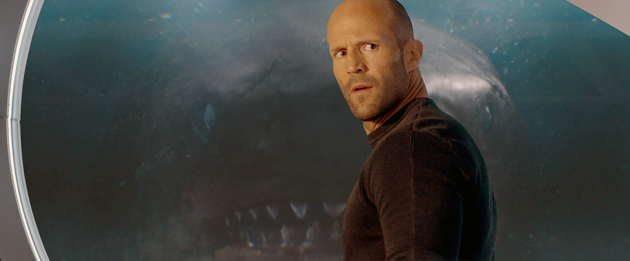 Jason Statham stands in front of a large underwater creature's face inside a research facility, appearing astonished