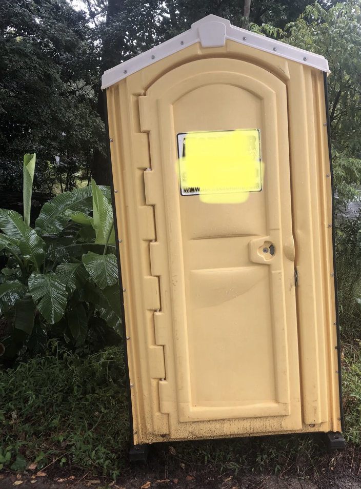 A yellow portable toilet is placed outdoors among green plants. The door is closed, and there is a small, unclear information sign on it