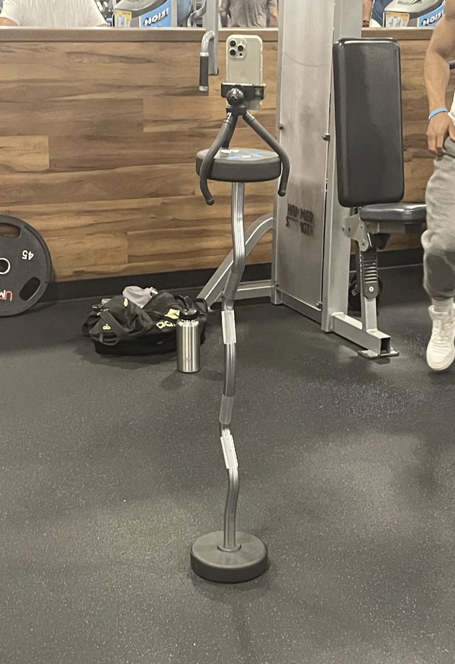 Phone mounted on a flexible stand at a gym, with gym equipment and a person partially visible in the background