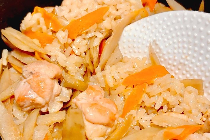 日本料理の炊き込みご飯が鍋に入っており、しゃもじが添えられている。鶏肉、ニンジン、ゴボウが混ぜられている。