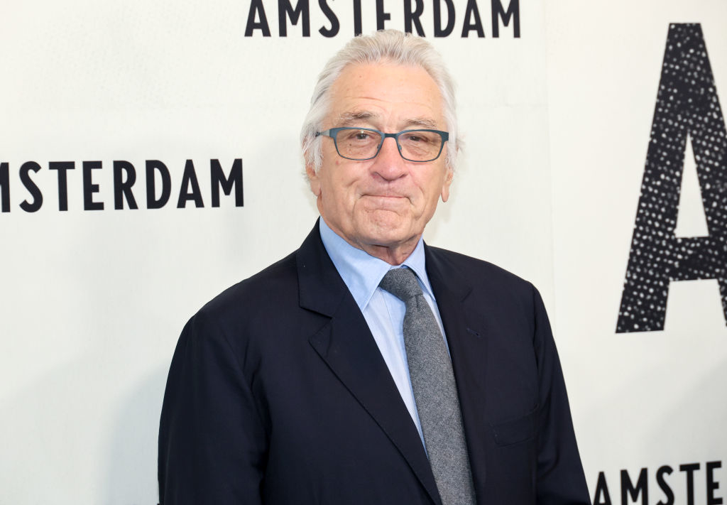 Robert De Niro at a premiere event for "Amsterdam" wearing a formal dark suit, light dress shirt, and tie, standing in front of a backdrop with the film's title