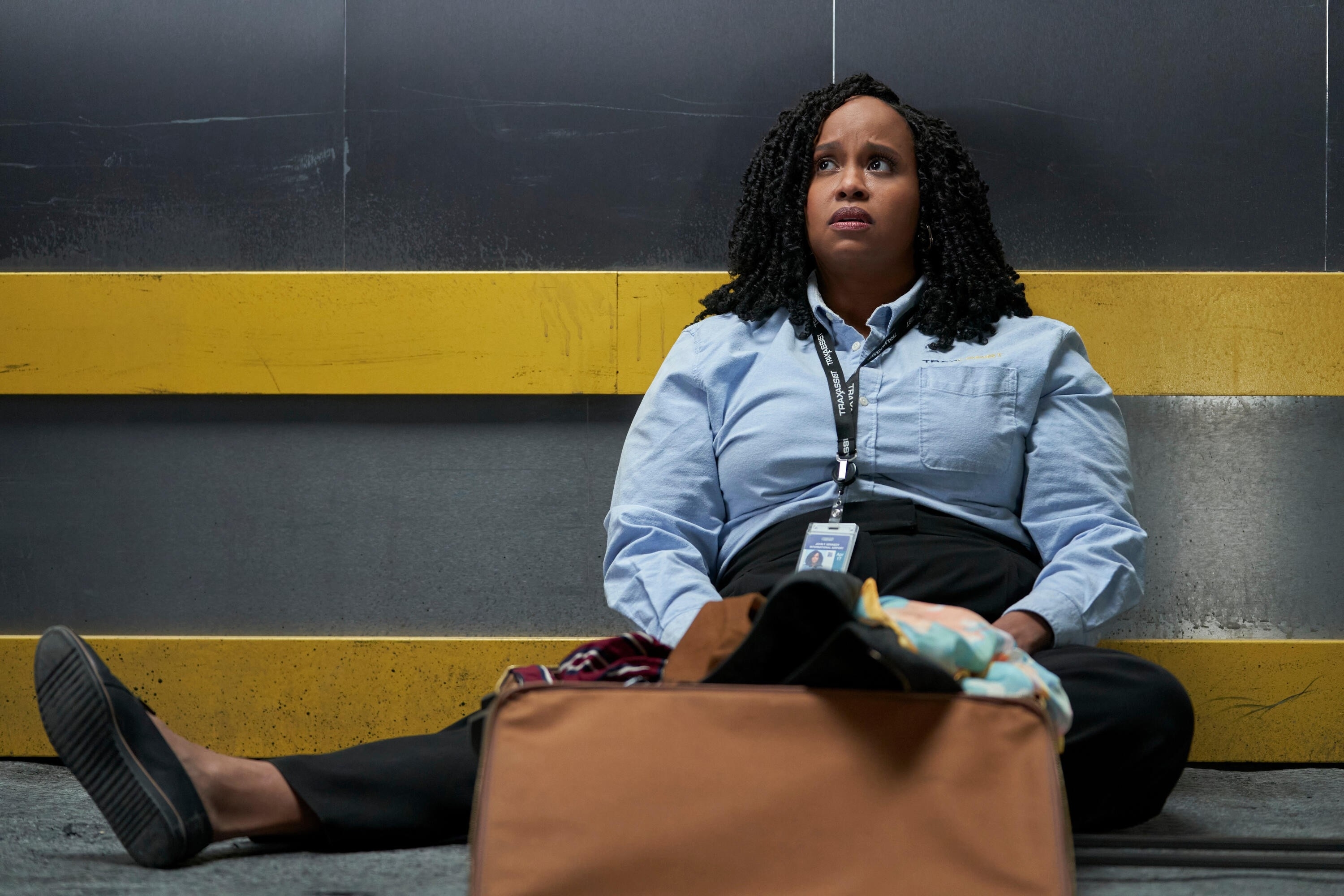 Natasha Rothwell in business casual attire sits on the floor against a wall next to a suitcase, looking up with a concerned expression in a a scene from How To Die Alone