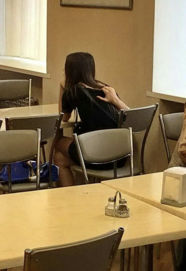 A person sits at a table with their back facing the camera in a casual indoor setting, while another person's hand reaches over to their shoulder