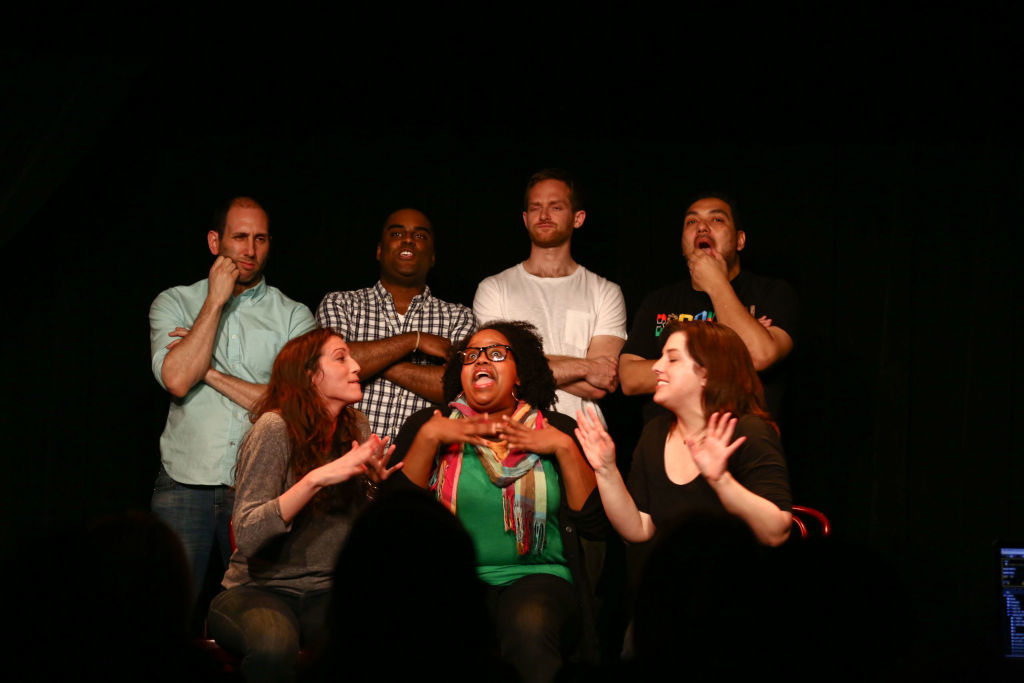 Six individuals, including Natasha, stand and sit on a stage, some with expressive gestures