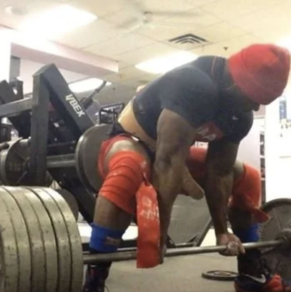 A muscular person is deadlifting heavy weights at a gym, wearing a beanie, a tight top, and shorts with knee wraps. Their intense focus is evident