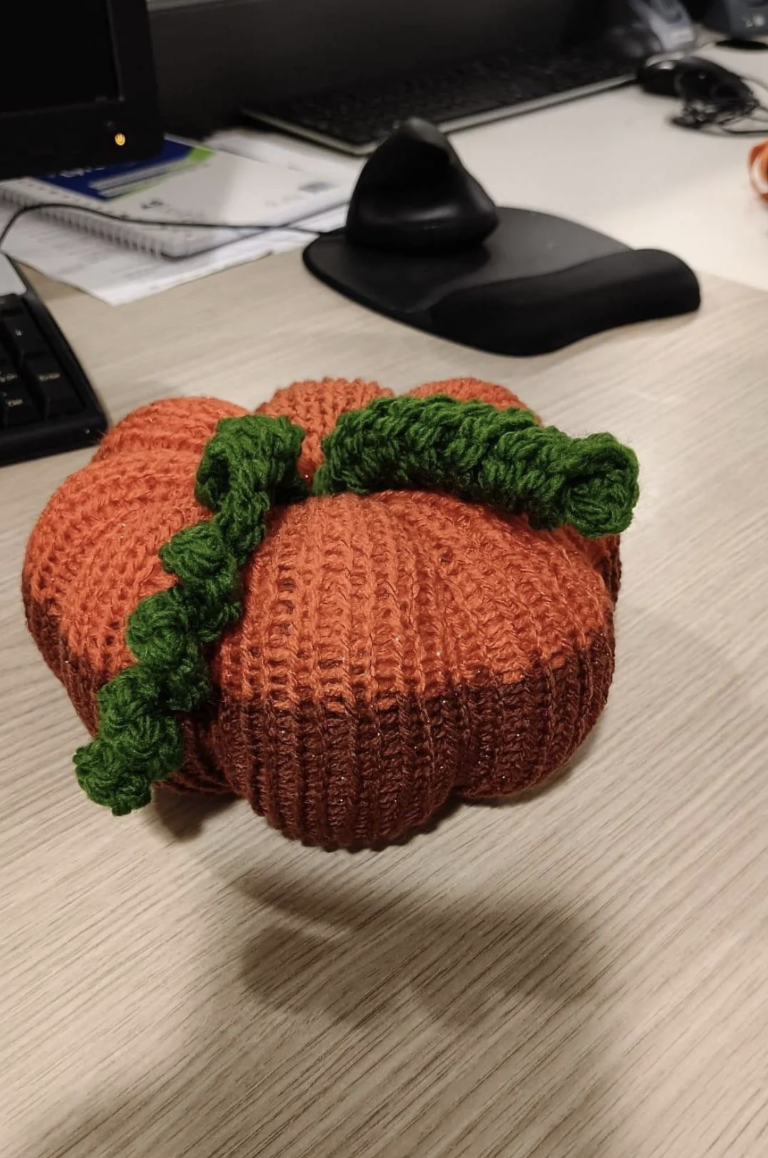 Knitted orange pumpkin with a green stem sitting on a desk next to office supplies