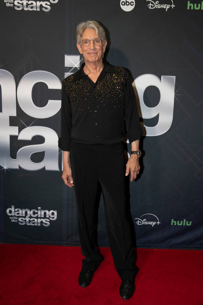 Eric Roberts standing on a red carpet in front of a "Dancing with the Stars" backdrop. He is wearing a black outfit with gold embellishments on the top