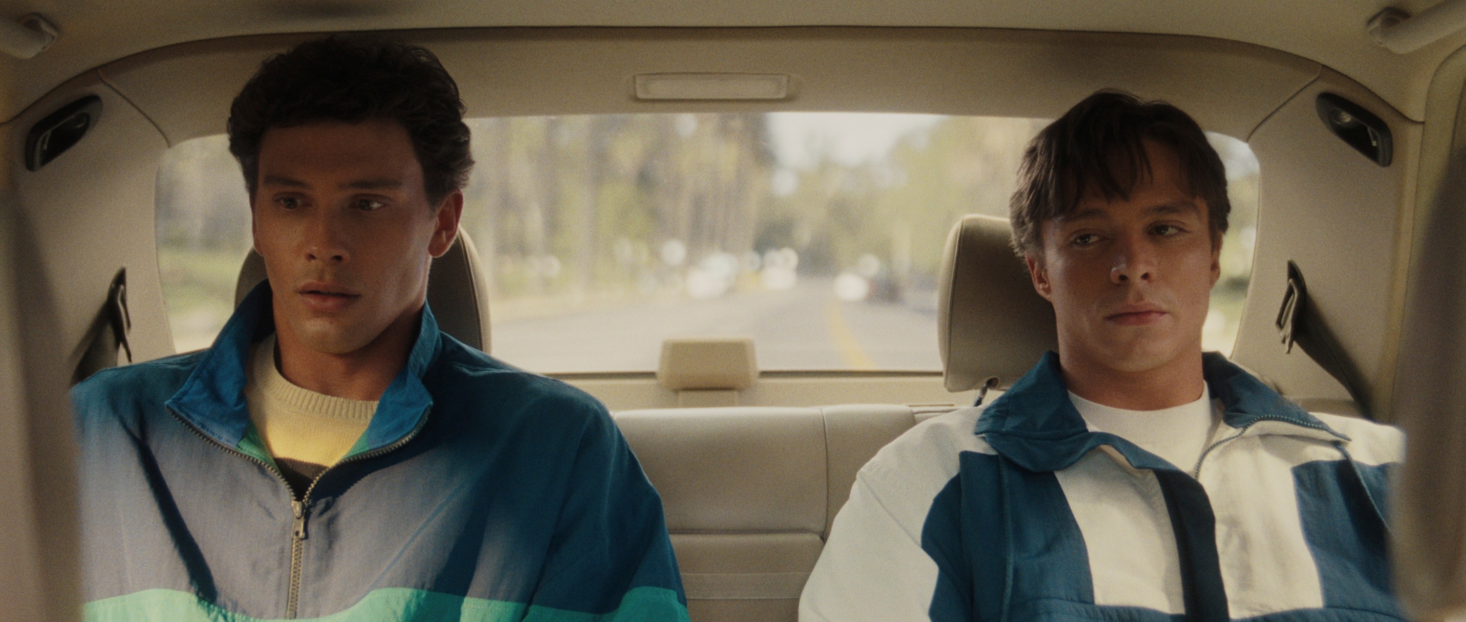 Two people, both wearing sports jackets, are sitting in the back seat of a car looking forward