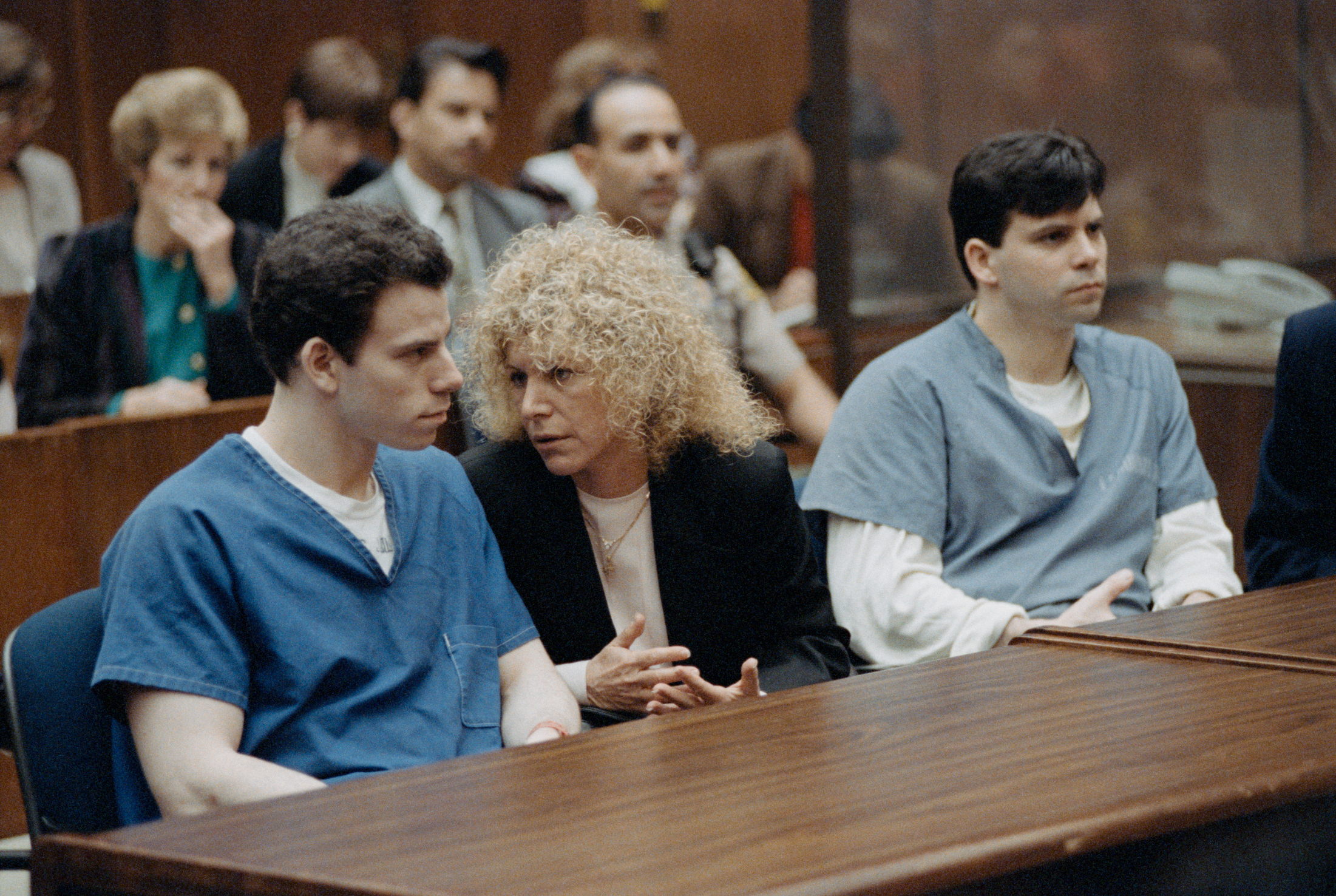 Erik Menendez (left) and Lyle Menendez (right) sit in a courtroom with an unidentified woman between them. They are all focused on a conversation