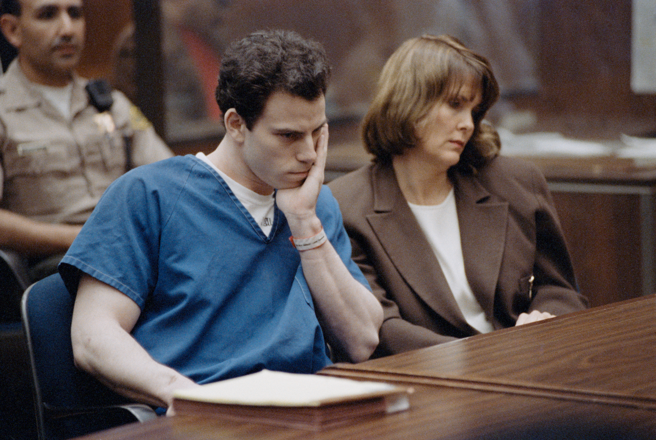 Man in blue prison uniform and woman in brown jacket sit in courtroom, looking contemplative and concerned