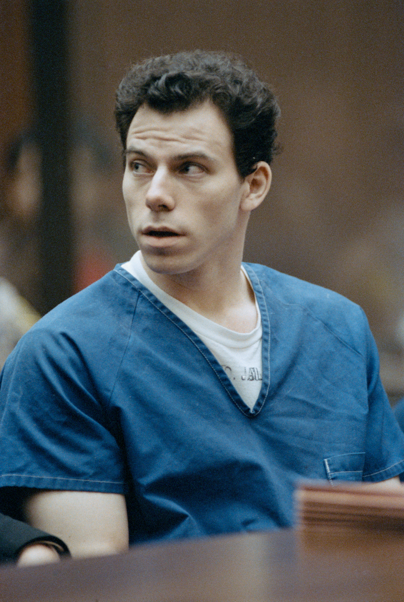 A person in a blue jumpsuit looks to the side while sitting in what appears to be a courtroom. The person has short, dark hair