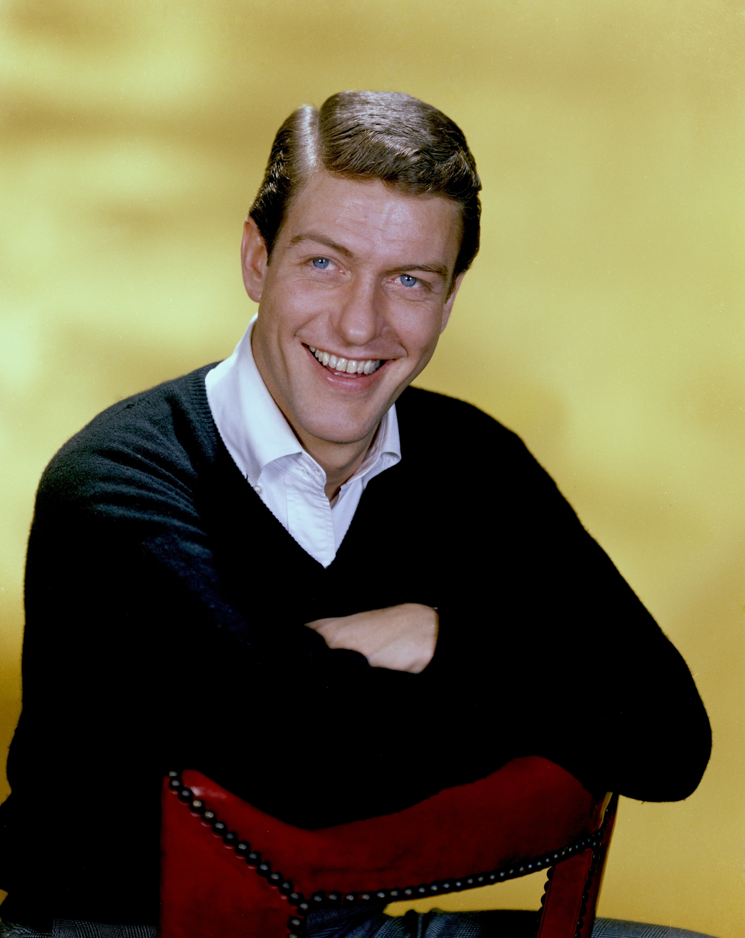 Dick Van Dyke smiling at the camera, leaning with arms folded on the back of a chair, wearing a white shirt under a black sweater