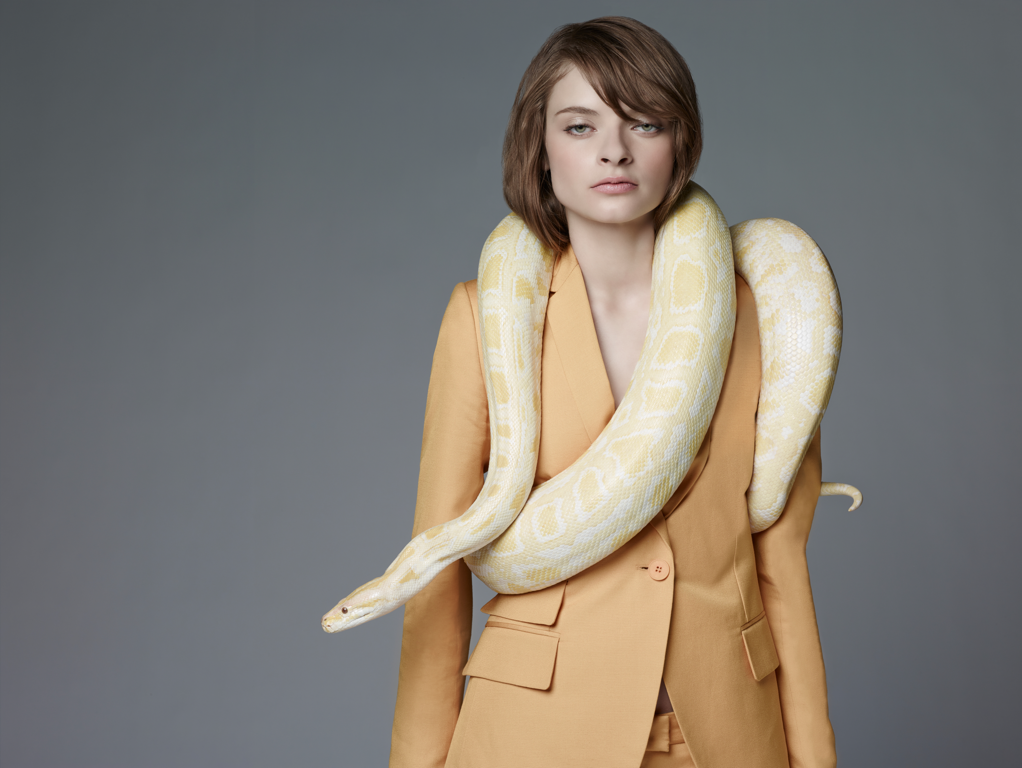 A woman in a tailored suit stands confidently with a large snake draped around her shoulders
