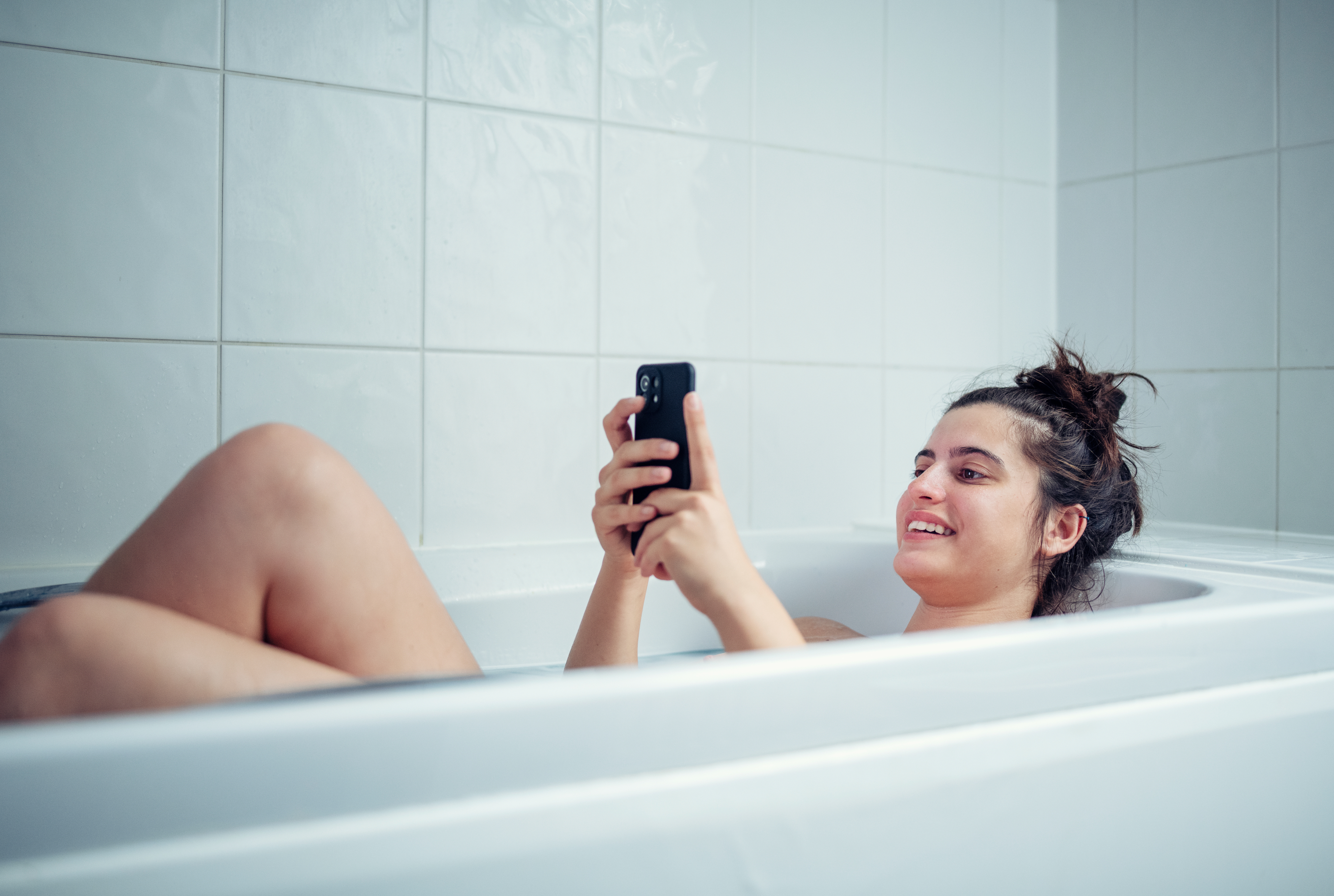 A smiling woman relaxing in a bathtub, holding and looking at a smartphone