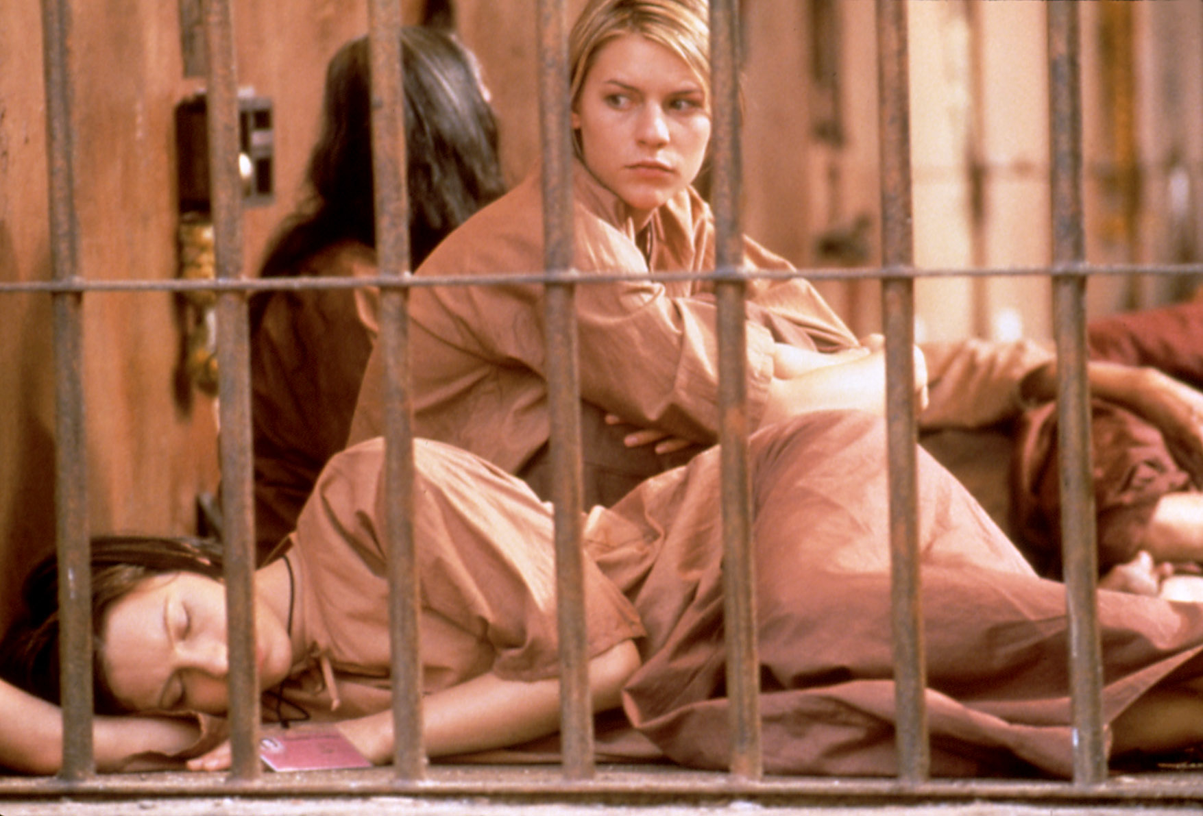 Claire Danes and another woman are behind bars. Claire is sitting and looking outside, while the other woman is lying on the ground sleeping. Both are wearing simple loose clothing