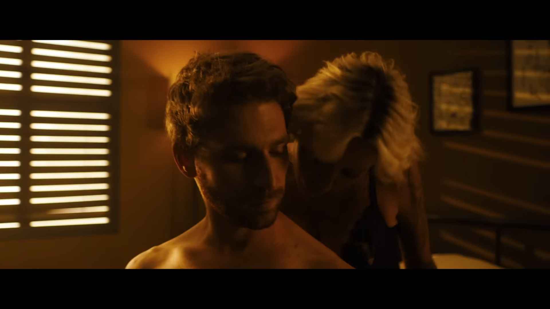 Shirtless man sits on bed while a woman in lingerie leans in close from behind in a dimly-lit room