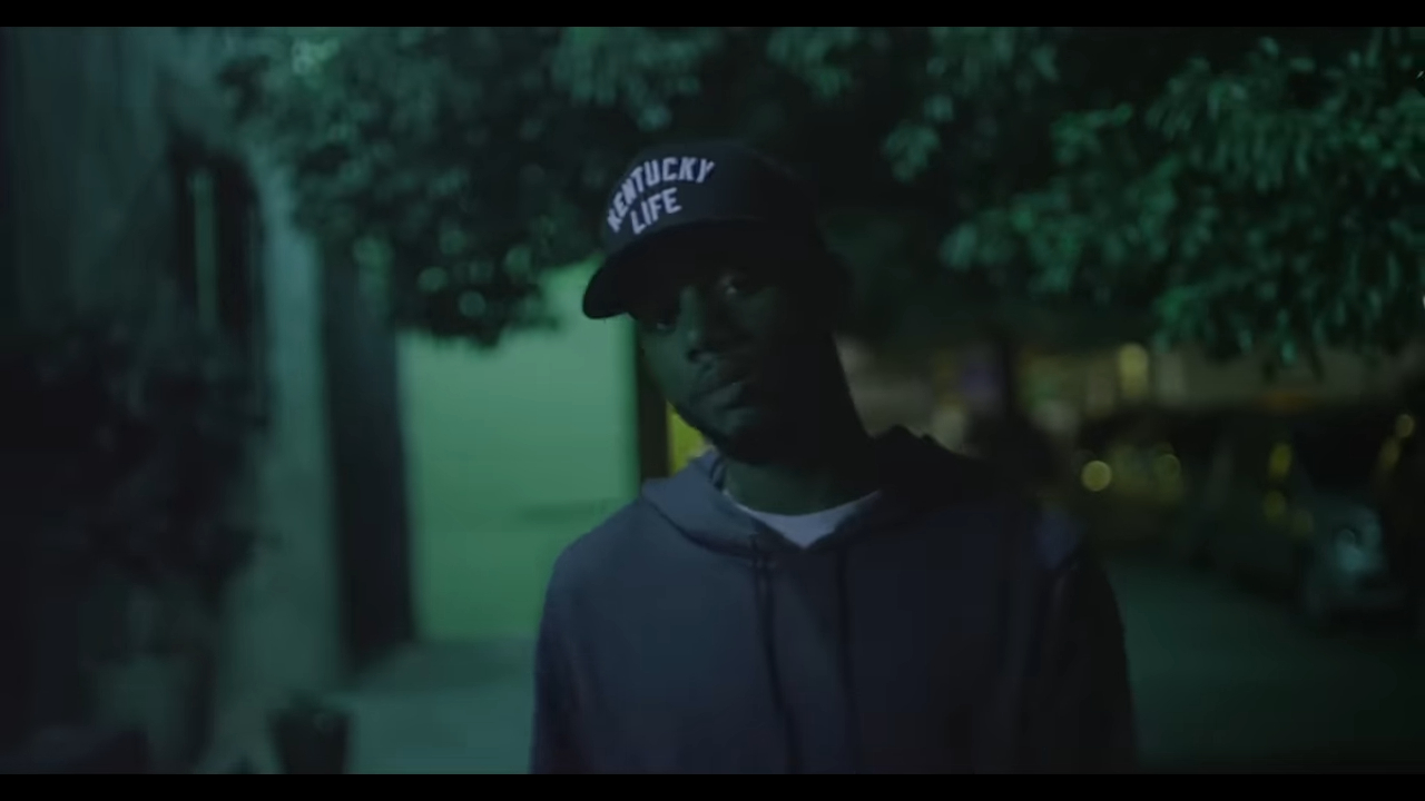 A man wearing a hoodie and a "Kentucky Life" hat stands on a dimly-lit street at night