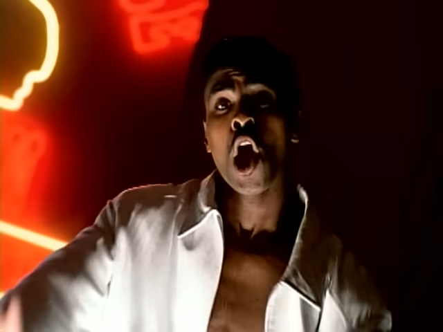 Ginuwine performing with a passionately expressive face, wearing an open white shirt. Neon signs glow in the background