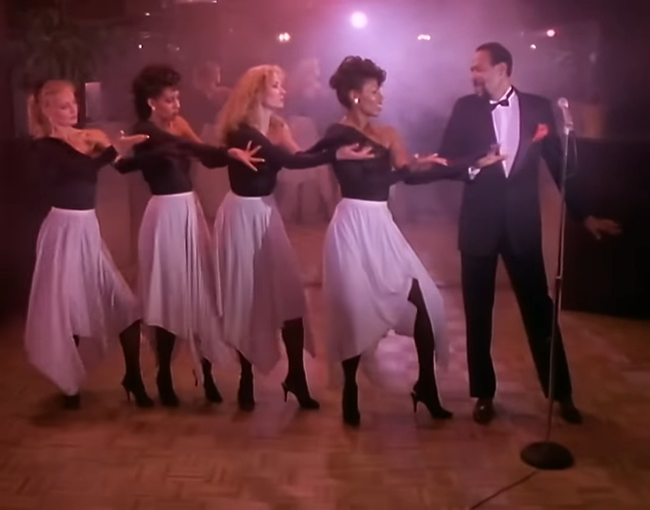Four women dressed in black tops and white skirts dance beside a man in a tuxedo and bow tie, performing on a stage with a microphone