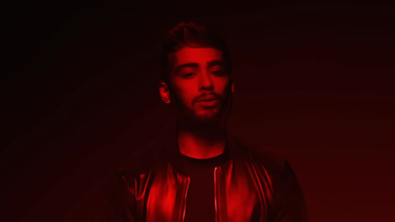Zayn Malik in a dark, dramatic setting, wearing a shiny jacket, poses against a shadowy background illuminated by red light