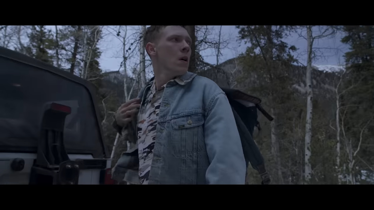 A man stands beside a vehicle in a forested area, looking startled and holding a backpack over his shoulder. Snow-dusted mountains are visible in the background