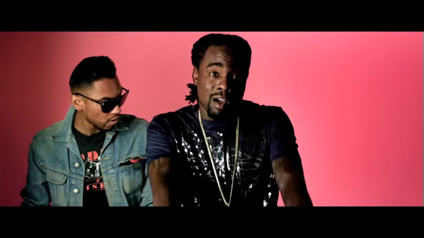 Miguel and Wale in a music video scene, with Miguel wearing a denim jacket and sunglasses and Wale in a black shirt with chain necklaces